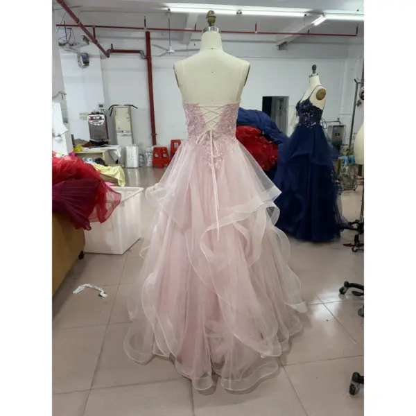 Back view of the Ophelia in Baby Pink sparkle tulle ball gown showcasing a delicate lace-up corset closure with criss-cross ribbons, intricate beaded and embroidered appliques on the bodice, layered tulle construction and cascading tiered ruffles creating a romantic sweep train.