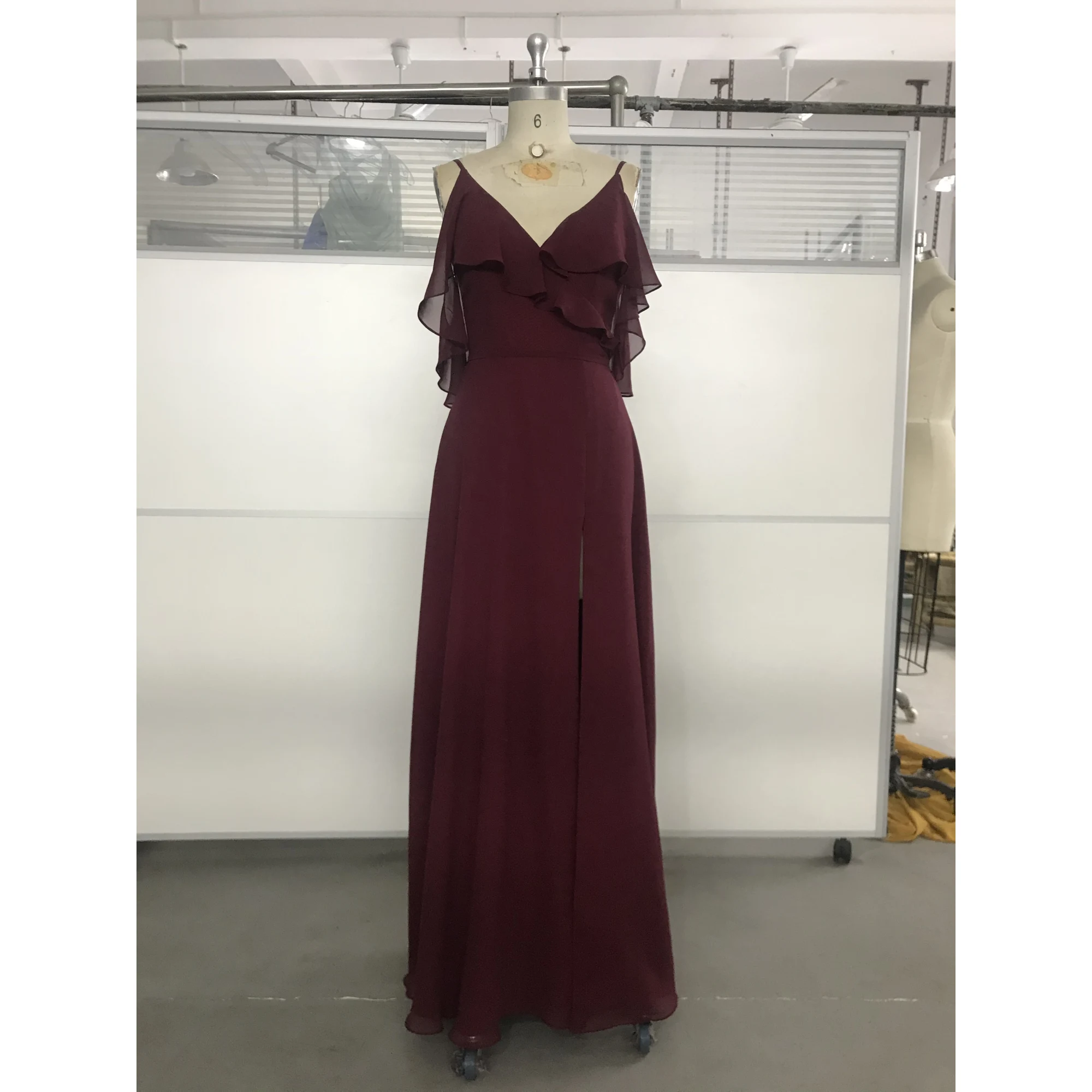 Elegant Capucine gown in rich Wine color featuring flowing Chiffon fabric, a deep V-neckline with cascading ruffles on the shoulders, and a sophisticated high slit that reveals graceful movement.