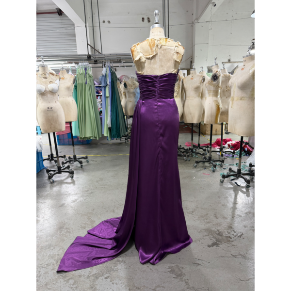Back view of the Laurence Eggplant Stretch Satin Ruched Mermaid Gown on a mannequin in a workshop setting, showcasing the sleek closed back with ruched detailing through the waist and hips, smooth stretch satin in deep eggplant purple that flows into a long sweeping train.