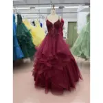 Stunning Crystal Burgundy tulle ball gown on a mannequin featuring a sweetheart neckline with intricate burgundy lace appliques, beading and sequins, fitted bodice and dramatic multi-tiered ruffled sparkling tulle skirt in a prom dress factory setting.